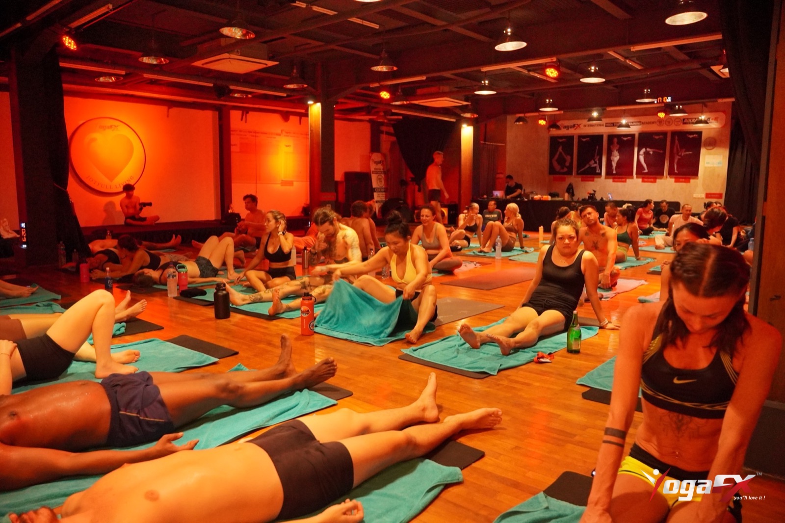 Rows of students lying between floor postures in a dimly lit heated studio during the recovery periods of class