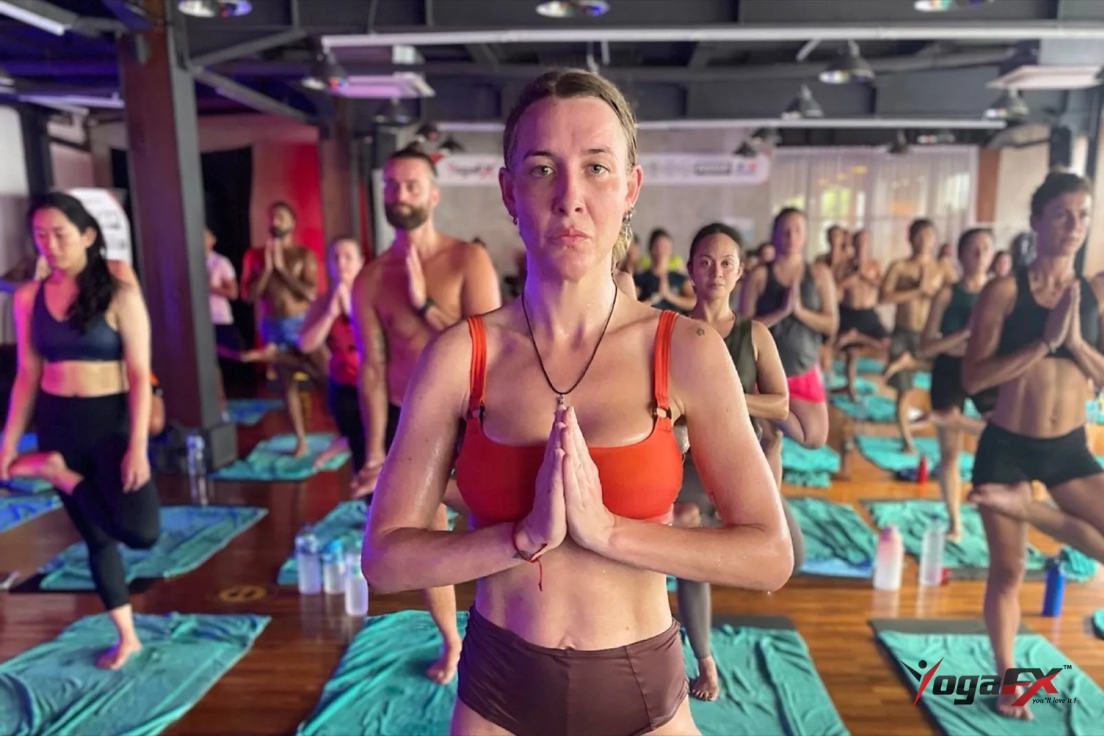 Student in prayer position at the front of a full hot yoga room before the balance series, showing the focus yoga championship athletes train